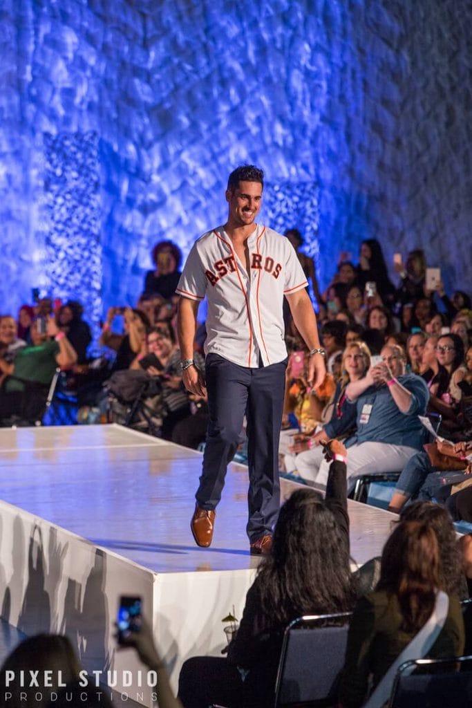 Male model walking on runway during fashion show in front of audience.