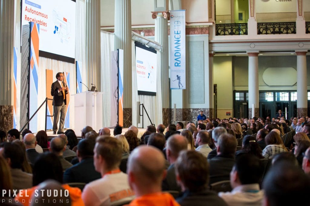 Speaker presenting at a professional conference with audience in a grand hall.