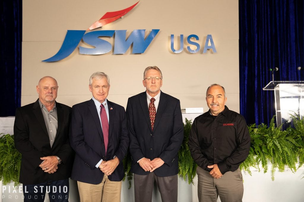 Group of four men at JSW USA corporate event, standing in front of logo.