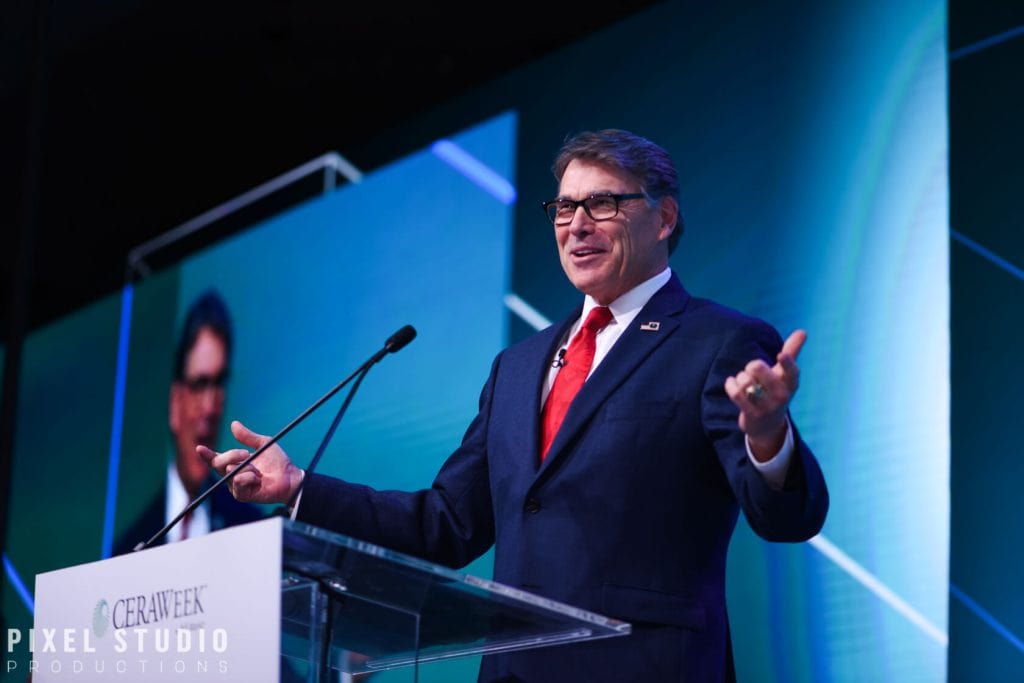 Rick Perry delivering speech at professional event with large screen behind him.