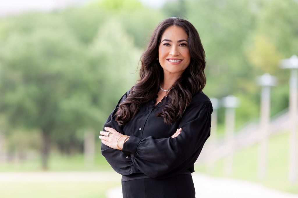 Outdoor professional headshot of a smiling woman with long dark hair in a black blouse, arms crossed, Houston