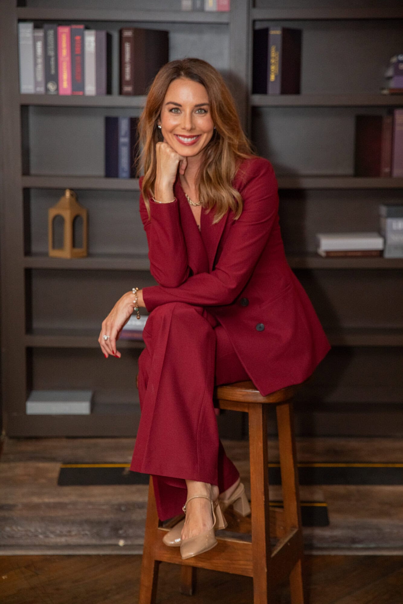 Confident woman in red suit sitting on a wooden stool in a modern library.
